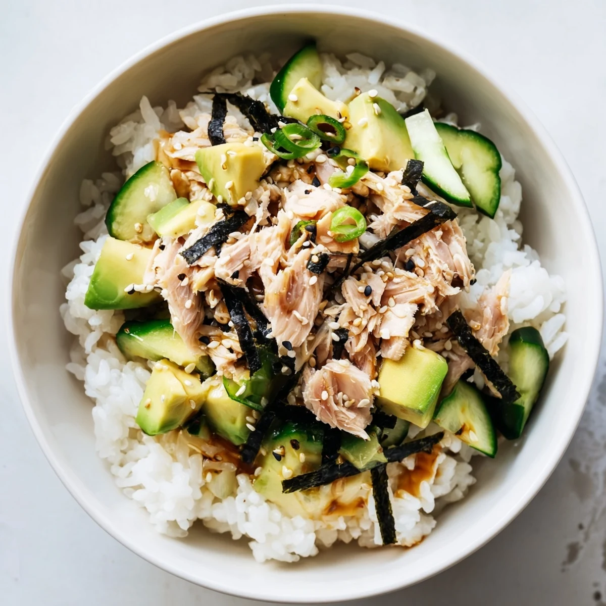 Fragrant jasmine rice topped with flaked tuna, diced avocado, cucumber, and scallions, drizzled with a savory ginger-sesame dressing in a white bowl.  