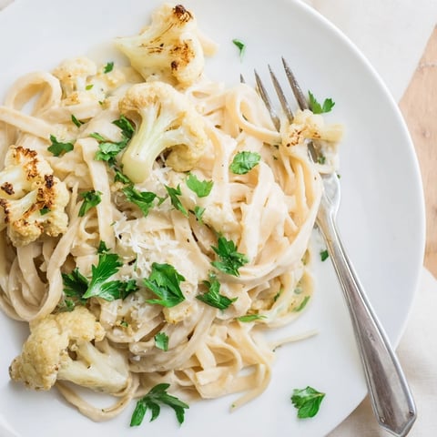 Golden-brown roasted cauliflower florets and garlic rest beside a bowl of creamy Roasted Cauliflower Alfredo sauce, ready to blend.