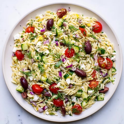 Greek Orzo Salad served in a white bowl with vibrant cherry tomatoes, crisp cucumber, and crumbled feta cheese, drizzled with a zesty lemon-oregano vinaigrette.