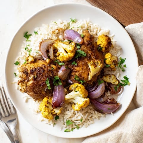 A single sheet pan holds seasoned chicken and caramelized cauliflower, garnished with fresh parsley and lemon wedges.  