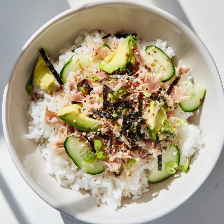 A close-up view of a vibrant Tuna Avocado Rice Bowl garnished with toasted sesame seeds, nori strips, and fresh cilantro on a wooden table.  